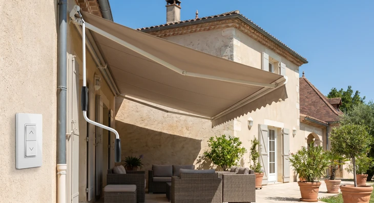 Photographie professionnelle d'une terrasse résidentielle montrant un store banne entièrement déployé en position ouverte. Au premier plan ou au centre de l'image, mettre en évidence le mécanisme de commande : soit une manivelle manuelle visible et accessible, soit un élément de motorisation (boîtier de commande sans fil, bouton mural). La terrasse est ensoleillée sous ciel bleu, le store banne crée une zone d'ombrage nette sur la terrasse. On peut voir discrètement du mobilier de terrasse ou des plantes pour montrer l'utilisation. L'arrière-plan montre une maison résidentielle classique. La photo doit clairement illustrer que le store peut être actionné manuellement ou de manière motorisée, en mettant l'accent sur la facilité d'utilisation et l'accessibilité de la commande.