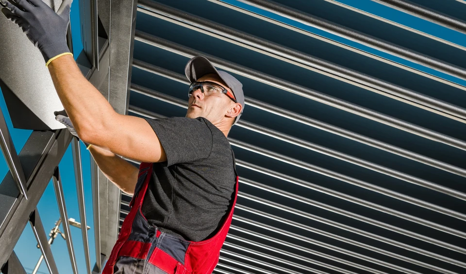 Ouvrier en tenue de travail rouge et noir installe des stores métalliques extérieurs sur façade de bâtiment
