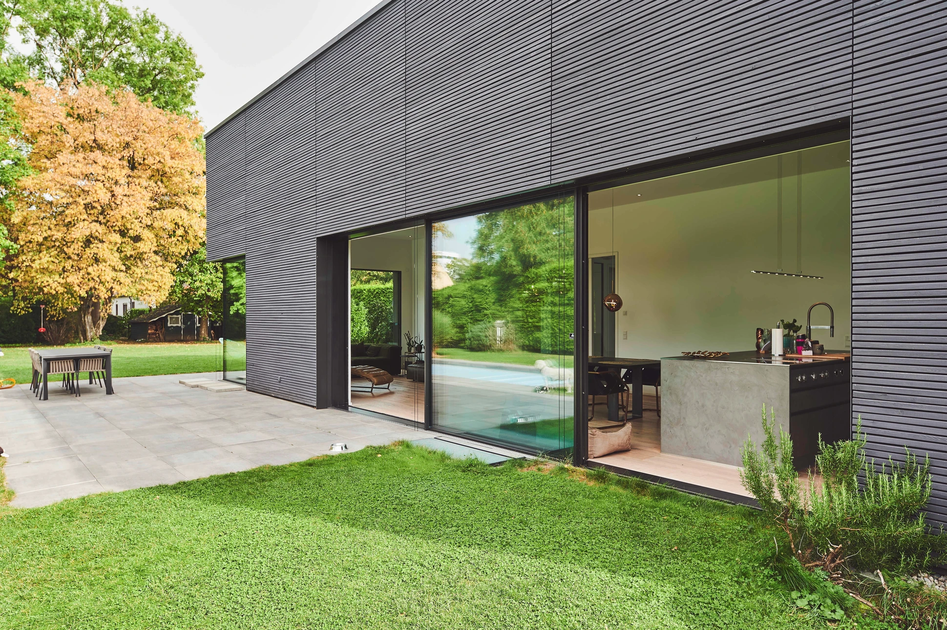 Photographie d'une grande baie vitrée ou façade vitrée d'une maison résidentielle sur laquelle est appliqué un film solaire transparent. La lumière naturelle pénètre encore abondamment la pièce intérieure visible derrière le verre (mobilier, espace de vie bien éclairé), tandis que la teinte légèrement tamisée du film et les reflets miroir partiels du film anti-chaleur sont visibles de l'extérieur. L'image doit montrer le contraste clair entre le verre traité (zone inférieure ou partielle) et le verre non traité pour illustrer l'effet du film. Aucune personne n'apparaît dans la scène. La composition privilégie la clarté et la luminosité pour traduire le concept de "filtrer la chaleur, garder la luminosité".