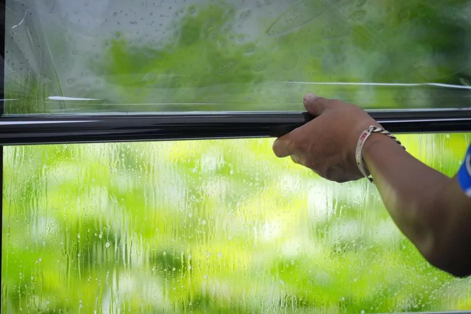 Gros plan d'une fenêtre ou baie vitrée ensoleillée avec un film solaire transparent appliqué sur le vitrage. La lumière naturelle passe clairement à travers le film, créant une légère teinte neutre. L'intérieur de la pièce derrière la vitre doit être visible, frais et lumineux, sans ombre marquée. Le film doit être appliqué de manière professionnelle et uniforme, mettant en évidence que la pose s'effectue sans modification de la menuiserie existante.